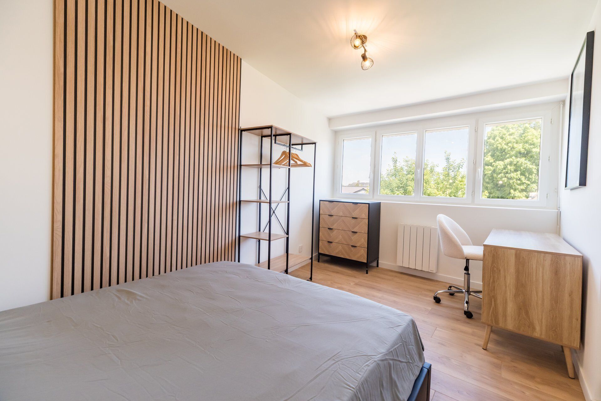 Une chambre à coucher comprenant un lit, un bureau, une chaise, un radiateur et une fenêtre donnant sur des arbres.