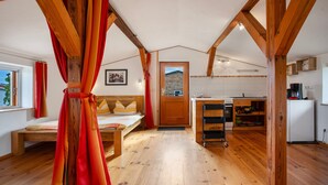 A cozy room with a bed, a wooden ceiling, a kitchen area with a refrigerator, and a window with curtains. - Apartment "Galerie" – Cozy Retreat Near Water and Forest (Kröslin)