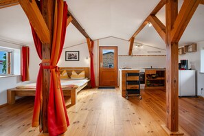 Ein gemütliches Zimmer mit einem Bett, Holzboden, einer Küchenzeile inklusive Kühlschrank und einem Fenster mit Vorhängen.
