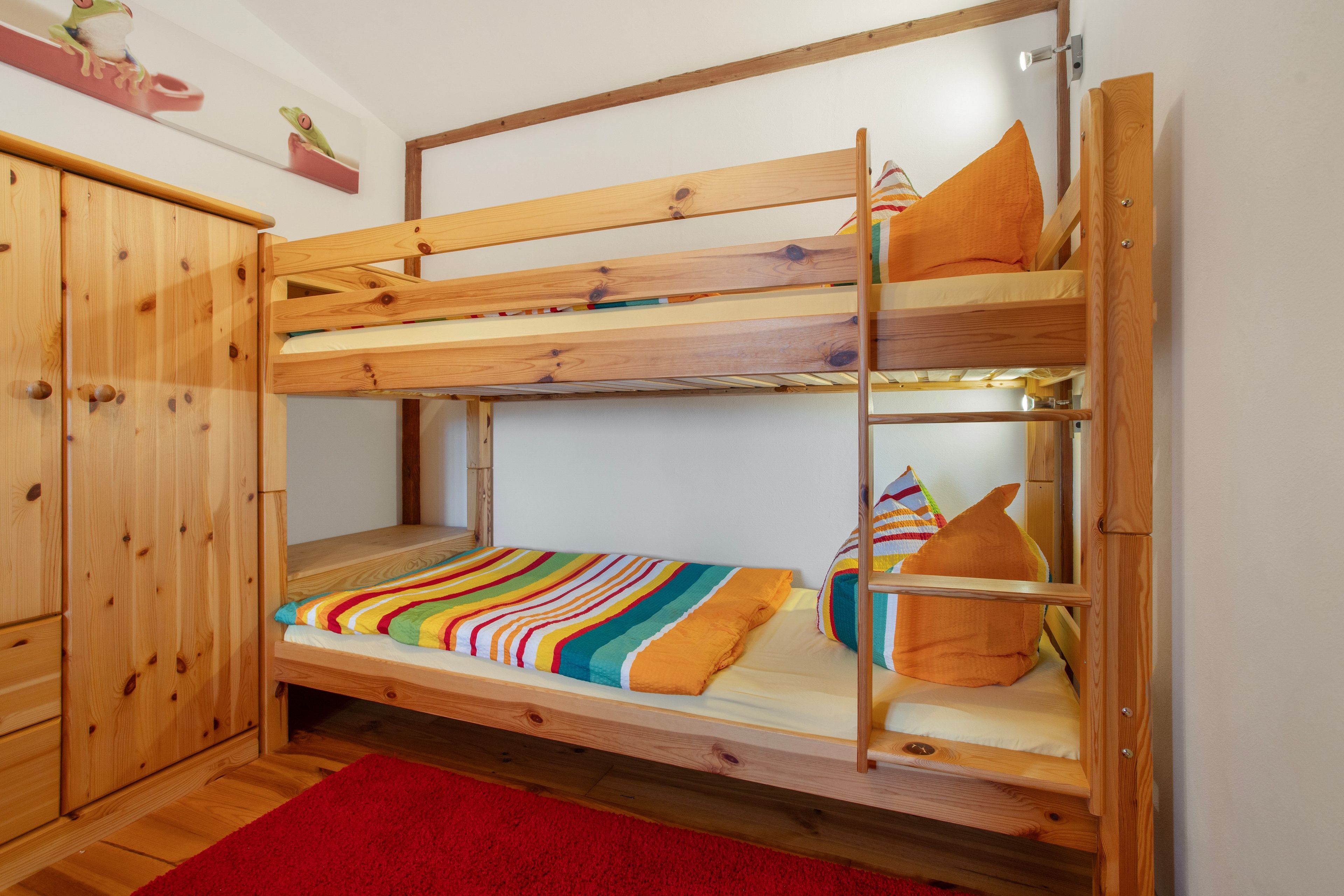 Una cama litera con estructura de madera, una alfombra roja y un pequeño armario.