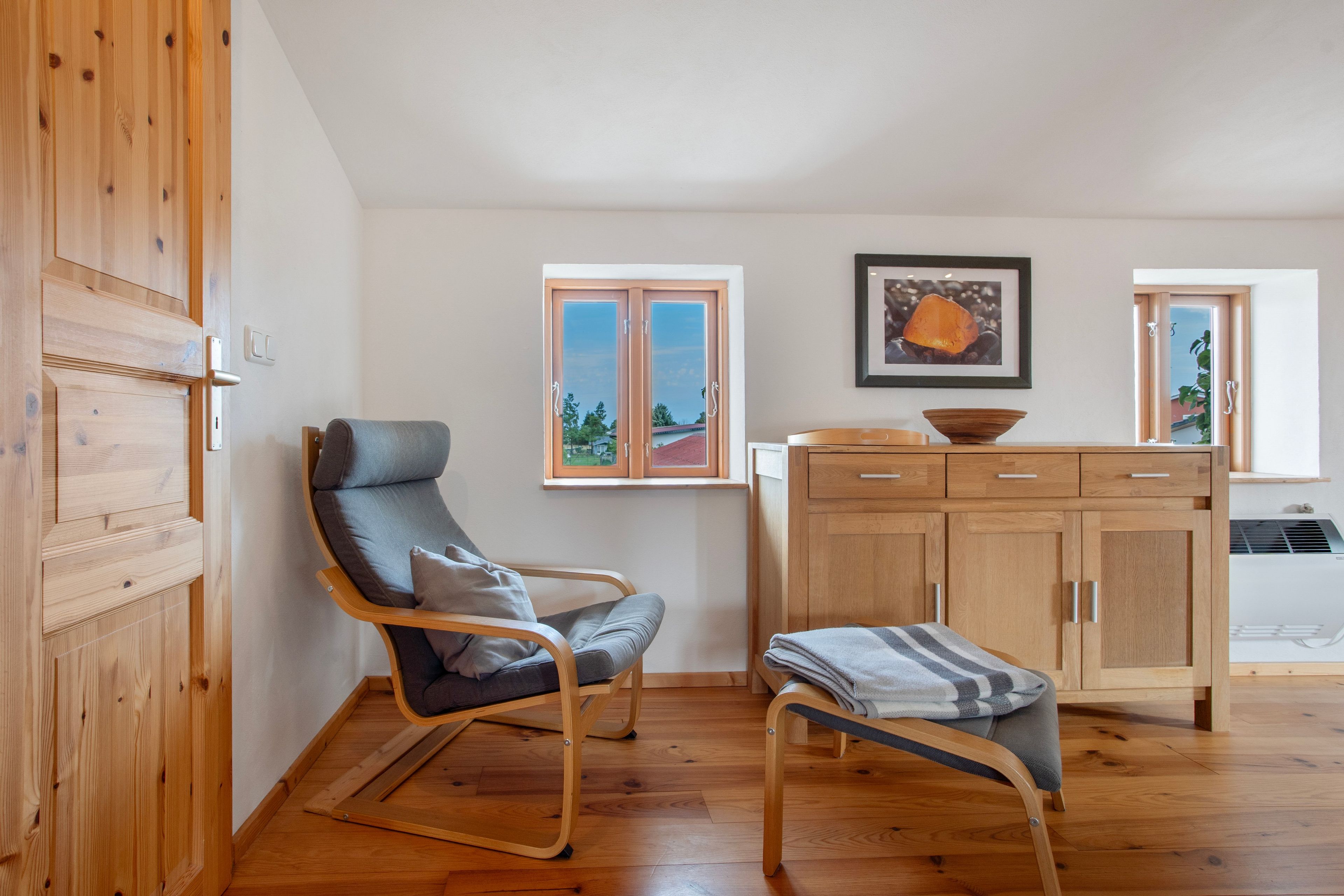 A room with a wooden floor, a grey upholstered chair, a wooden cabinet, and a framed picture on the wall.