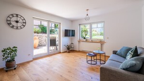 Een ruime woonkamer met een groot raam, een flatscreen televisie, een houten vloer en een grijze bank.