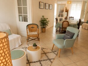 Ein Wohnzimmer mit einem Korbstuhl, einem blauen Sessel, einem kleinen Tisch mit einer Pflanze und einem Essbereich im Hintergrund.