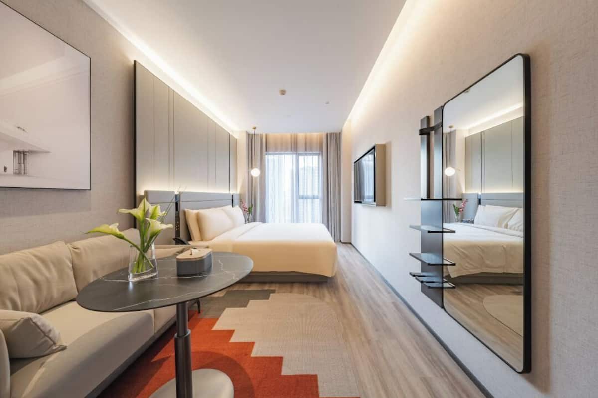 Una habitación de hotel moderna con cama, sofá, mesa de centro, espejo y un ventanal grande.