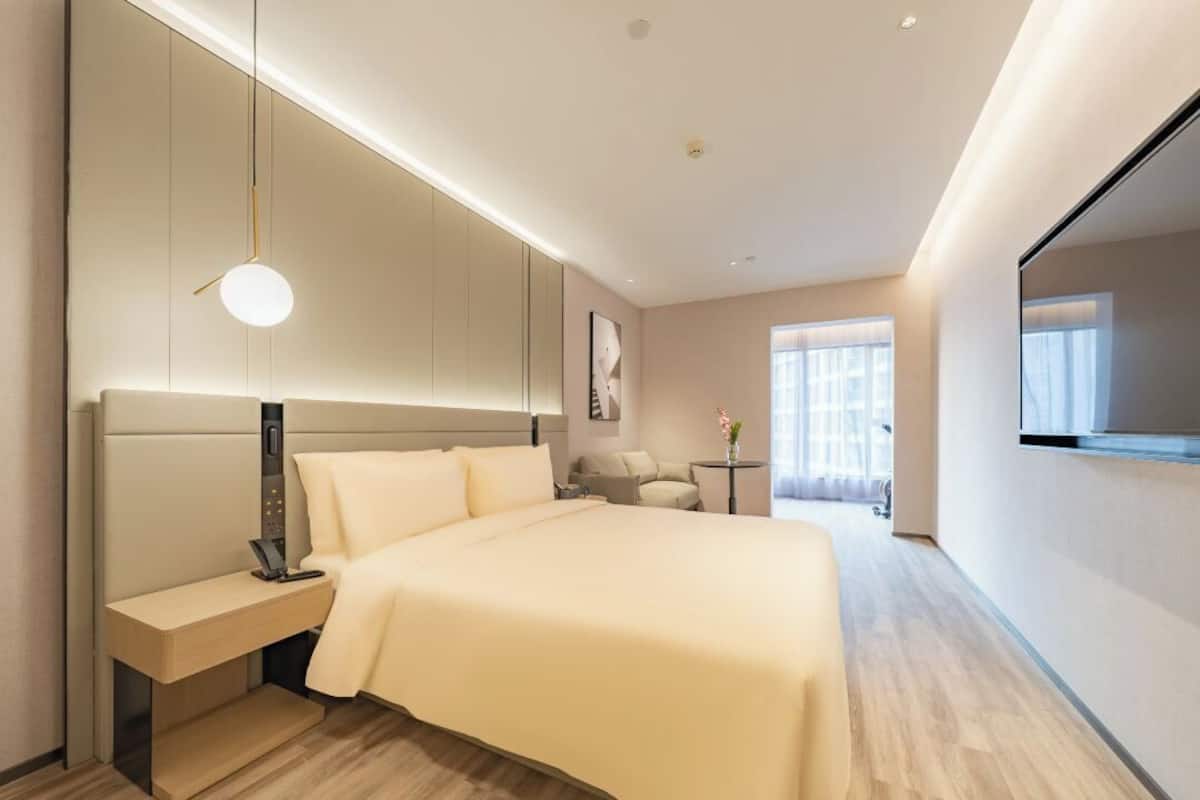 Una habitación de hotel moderna con una cama grande, una mesita de noche, un escritorio pequeño, una silla, un televisor montado en la pared y una ventana con cortinas.