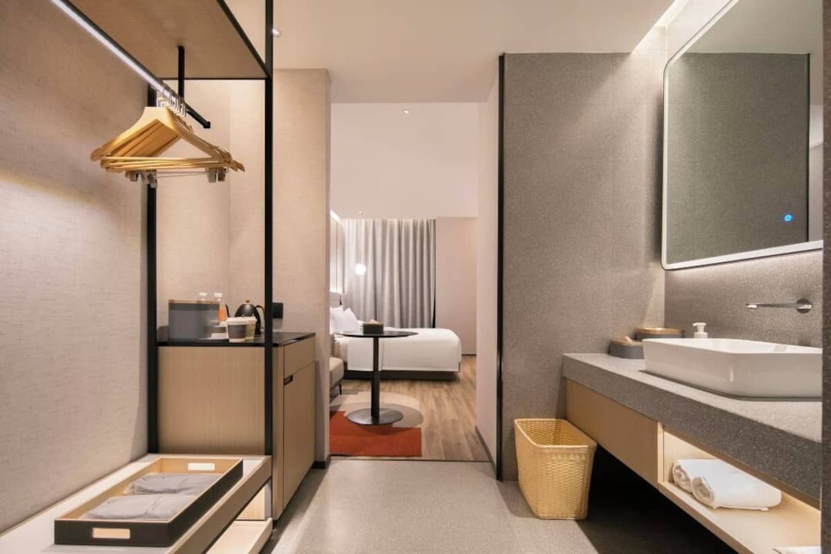 Un baño de hotel moderno con un espejo grande, un lavamanos, un perchero para toallas, una mesita pequeña y una cama visible en la habitación contigua.