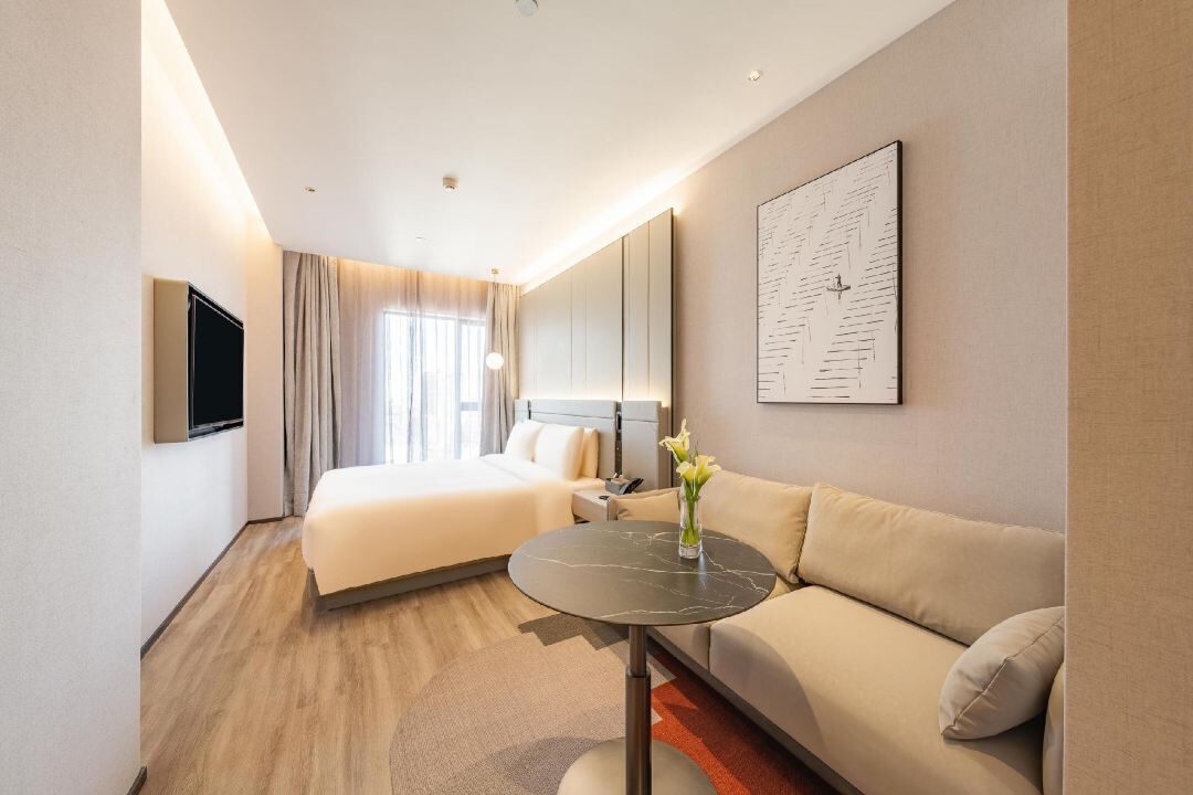 A hotel room with a bed, a sofa, a TV, a painting, and a vase with flowers.