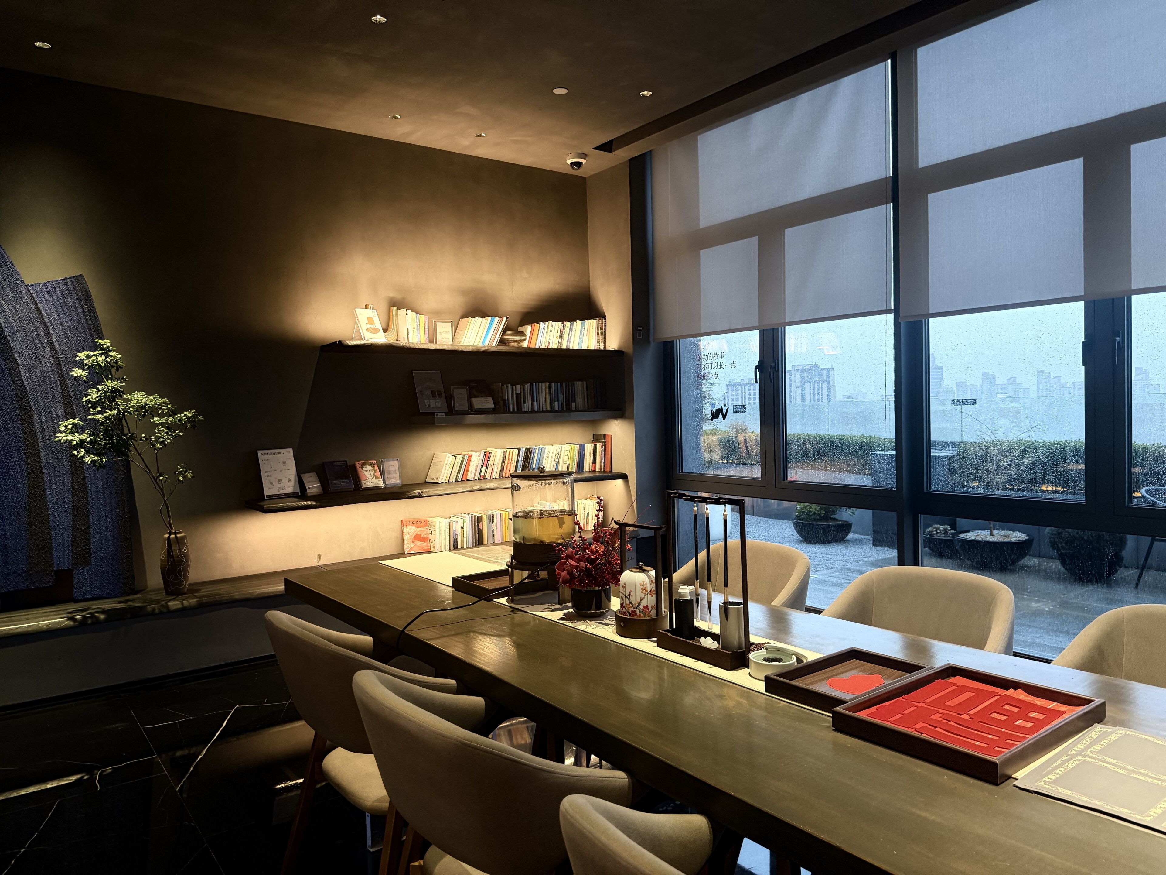 A modern conference room with a long table, chairs, a bookshelf, and a view of the city through large windows.