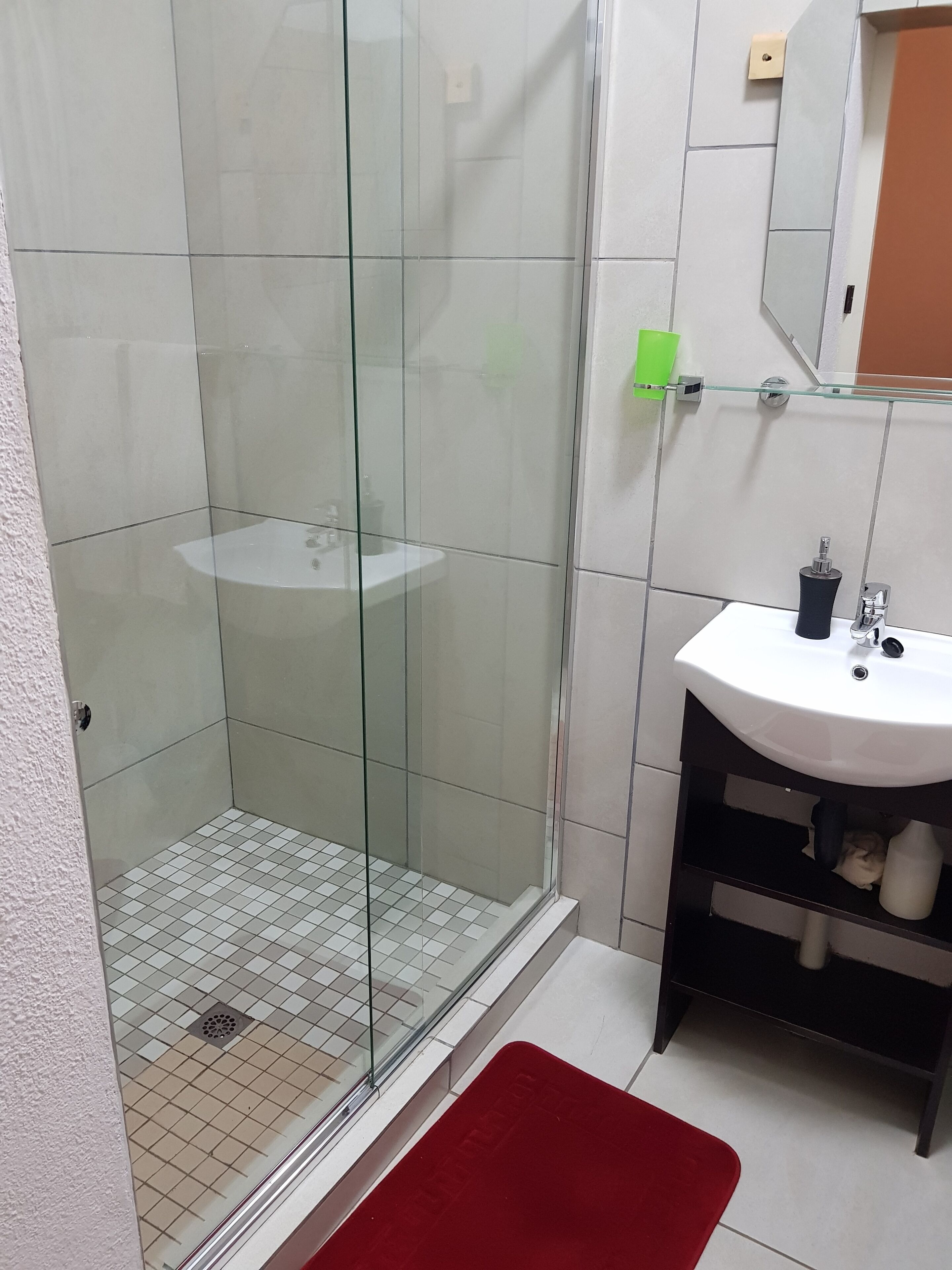 A bathroom with a glass shower enclosure, a white sink, and a red bath mat.