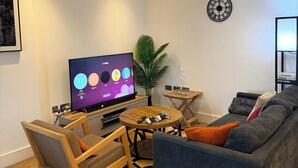 Moderni olohuone, jossa on litteänäyttöinen televisio, pelikonsoli, sohva, sohvapöytä ja huonekasvi.
