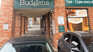 Rakennuksen edessä on kyltti, jossa lukee ”Budgens”, ja sen edessä on pysäköity auto. Toinen kyltti ilmoittaa, että liike on avoinna joka päivä klo 7–23.