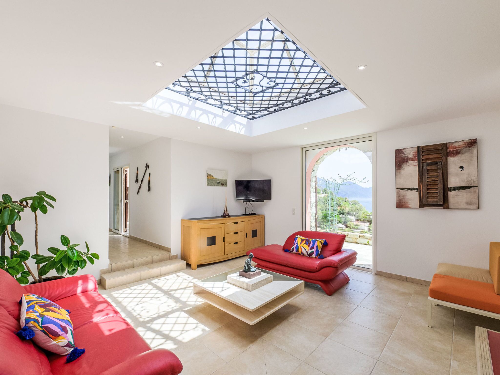 Ein geräumiges Wohnzimmer mit hoher Decke, einem Fernseher, einem roten Sofa, einer Holzkommode und Blick ins Freie.