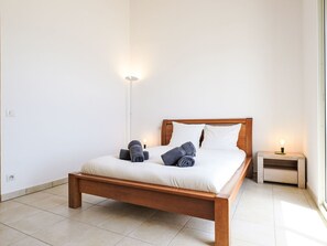 A neatly made bed with white linens and rolled towels, a wooden headboard, a bedside table with a lamp, and a tiled floor. - Kanelata by Interhome (Canari)
