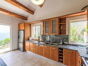 A modern kitchen with wooden cabinets, a stainless steel refrigerator, a granite countertop, and a view of the sea. - Kanelata by Interhome (Canari)