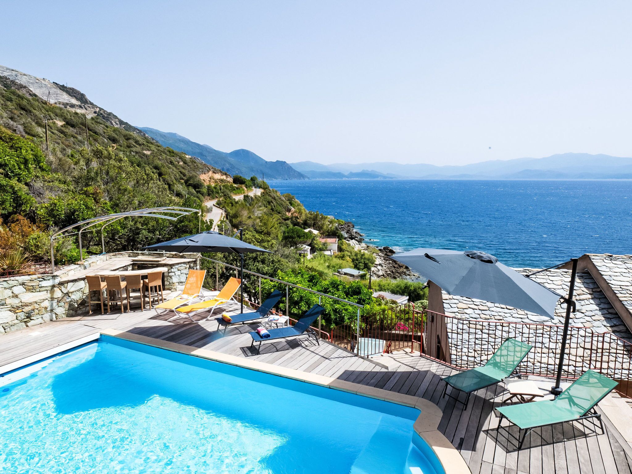 A swimming pool with lounge chairs and umbrellas, overlooking a coastal landscape with mountains and the sea.