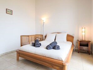 A neatly made bed with pillows and rolled towels, a wooden headboard, a bedside table with a lamp, and a small bedside drawer.