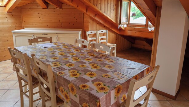 A dining area with a table set for four, wooden chairs, a window with a view, and a kitchen in the background. - Apartment 12 Maso Anna (Mastellina)