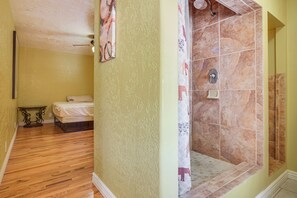 A room with a bed, a small table, a shower with a tiled wall, and a ceiling fan.