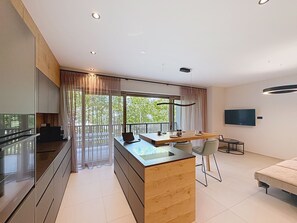 Eine moderne Küche mit einer Holztischinsel, einem Essbereich mit Tisch und Stühlen, einem an der Wand montierten Fernseher und einem Balkon mit Blick auf Bäume.