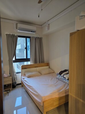 Un dormitorio con cama, unidad de aire acondicionado, ventana con cortinas, armario de madera y una mesita de noche.
