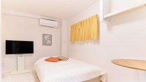 A bedroom with a bed, a flat-screen TV, an air conditioner, a window with yellow curtains, and a wooden side table. - Zenith Nishinakanobu 2 (Shinagawa)