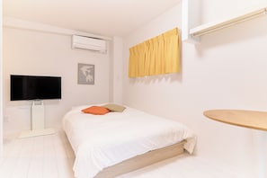 A bedroom with a bed, a flat-screen TV, an air conditioner, a window with yellow curtains, and a wooden side table. - Zenith Nishinakanobu 2 (Shinagawa)