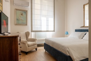 Ein Schlafzimmer mit einem Bett, einem Sessel, einer Holzkommode, einem Fernseher, einem Fenster mit Jalousien und einem Bild an der Wand.