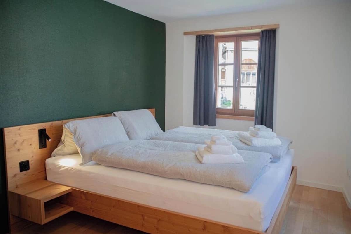 A room with a wooden bed, a nightstand, a window with curtains, and a view of buildings outside.