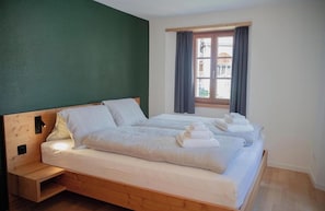 A room with a wooden bed, a nightstand, a window with curtains, and a view of buildings outside.