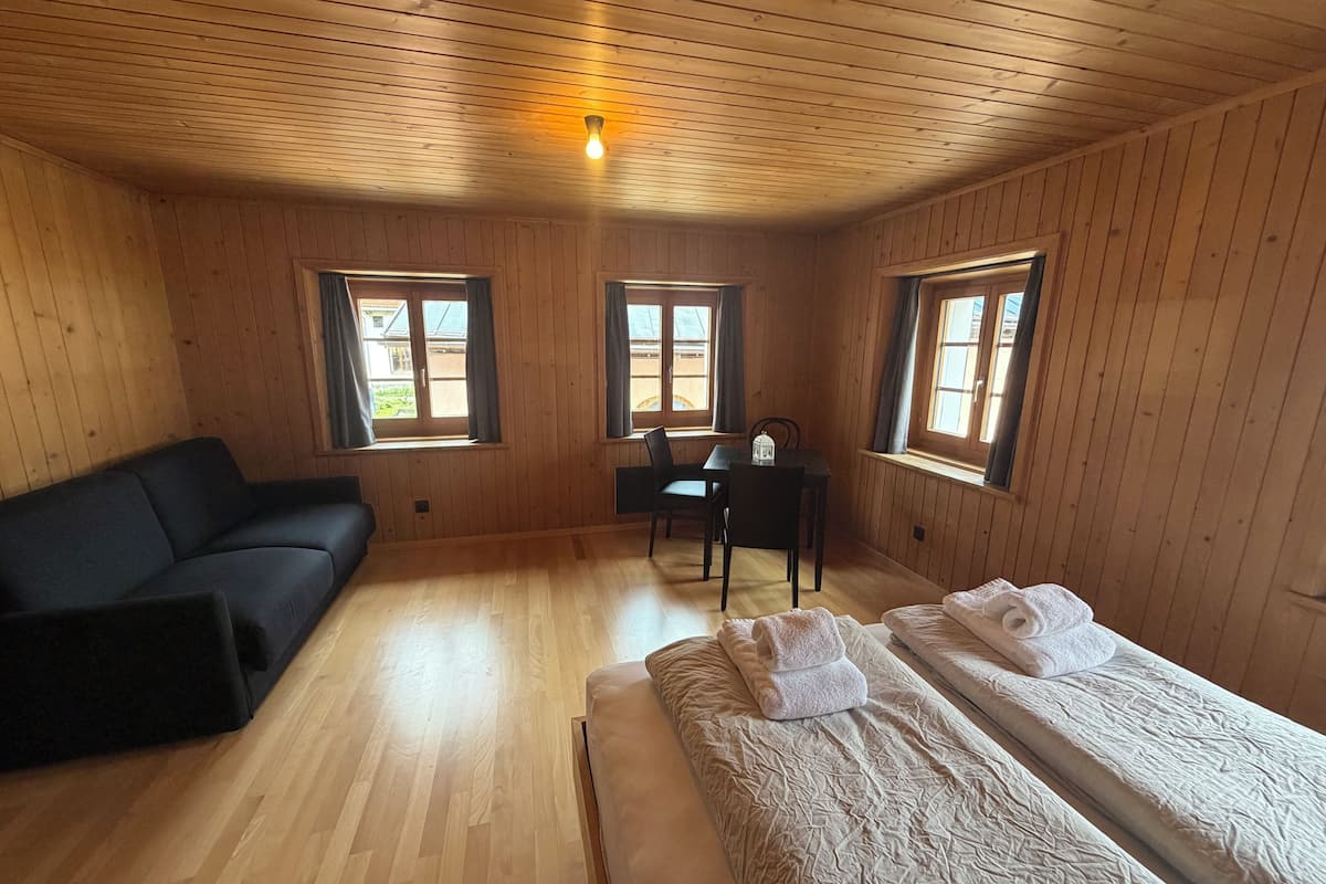 A wooden-paneled room with a sofa, two beds, a desk, and windows.