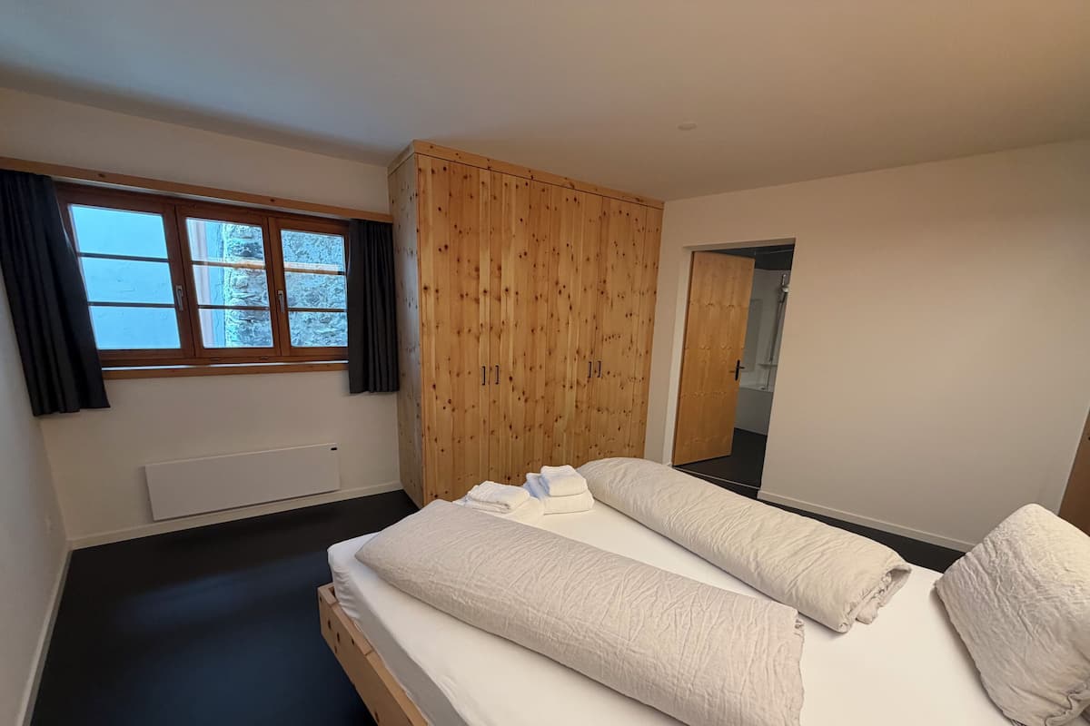 A bedroom with a wooden wardrobe, a window with curtains, a radiator, and a bed with pillows.