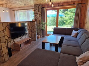 A modern living room with a flat-screen TV, stone wall, wooden ceiling, and a large window with a view of greenery.