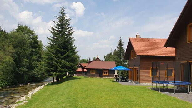 A wooden cabin with a red roof, a grassy yard, and a small stream. - Chalets Hana (Demänovská Dolina)