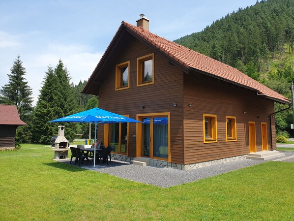 A wooden house with a red-tiled roof, a patio with outdoor seating, and a stone fireplace.
