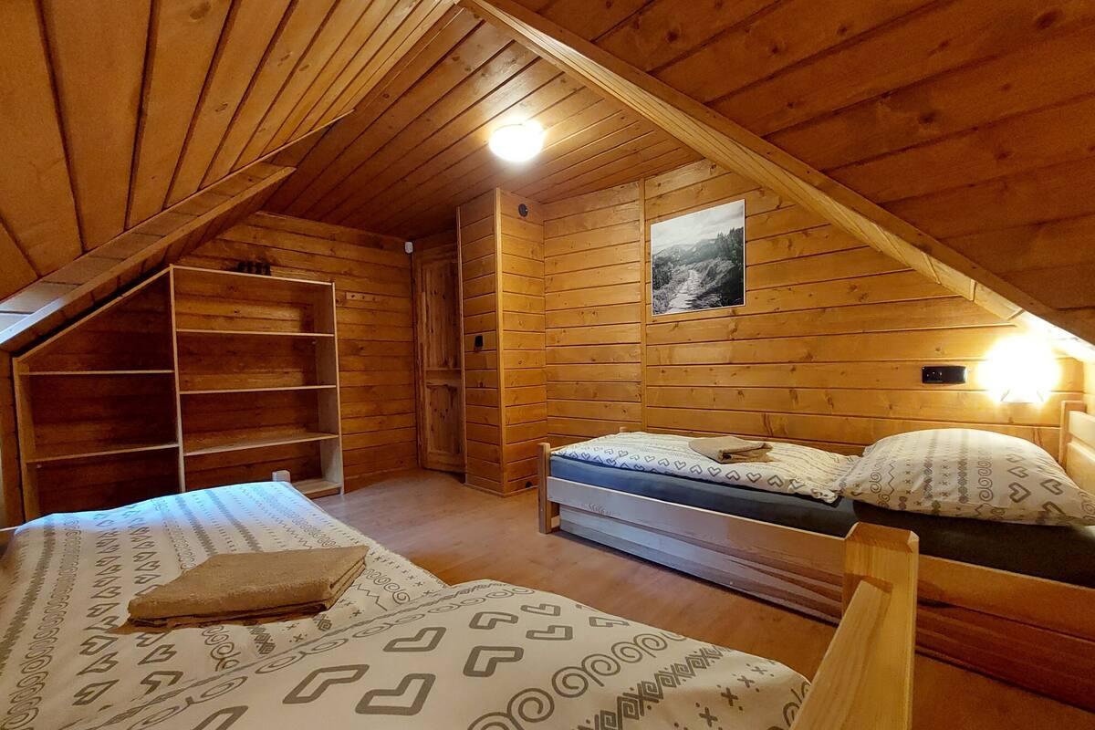 A wooden cabin bedroom with a sloped ceiling, a bed with patterned bedding, a nightstand, a shelf unit, and a wall-mounted light.
