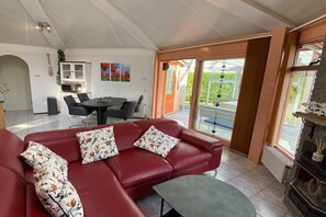 En rød lædersofa med blomsterpuder, et spisebord med stole, et køkken og udsigt gennem store glasdøre.