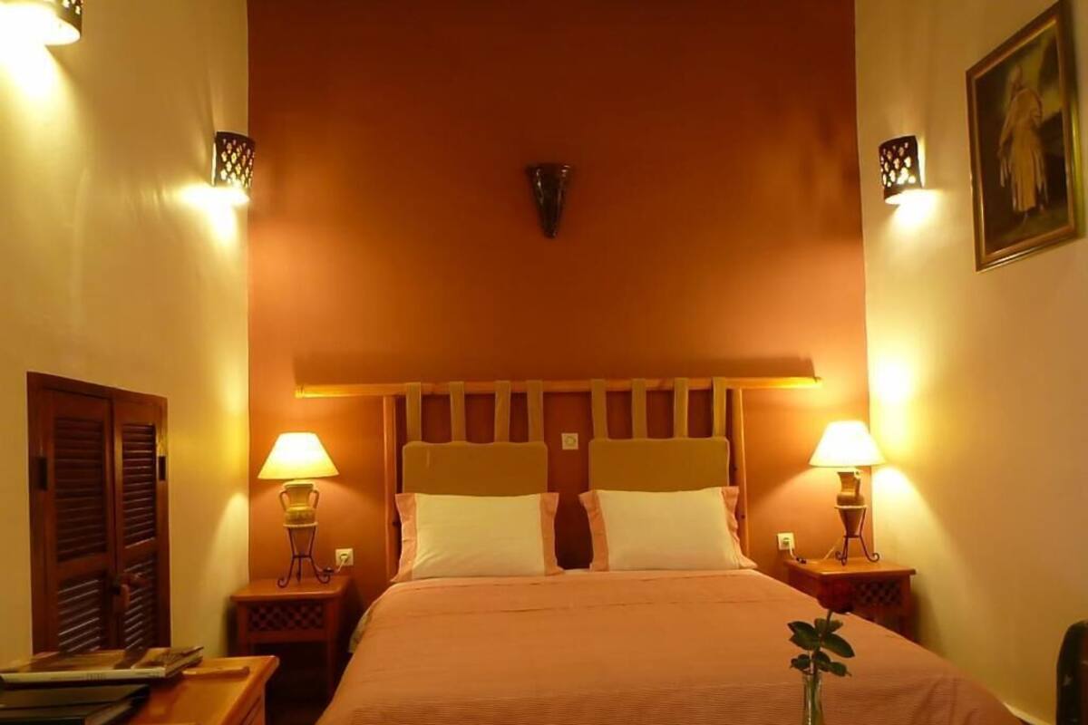 A hotel room with a bed, two bedside tables with lamps, a wooden headboard, and a wall-mounted light fixture.