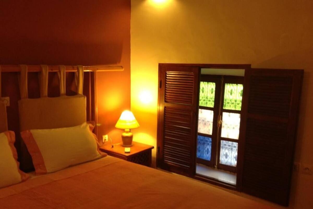 A hotel room with a bed, a nightstand with a lamp, a window with shutters, and a view of greenery outside.