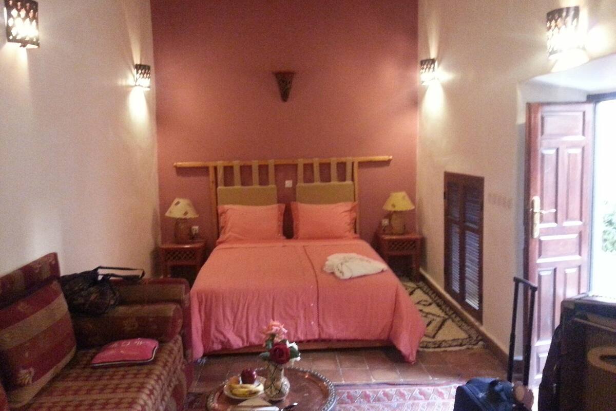 A hotel room with a bed, two bedside tables, a chair, a small table with a vase and a plate, and a door with a window.