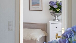 Um quarto com uma cama, uma mesa de cabeceira com um vaso de flores, um quadro na parede e uma porta que dá para outro cômodo.