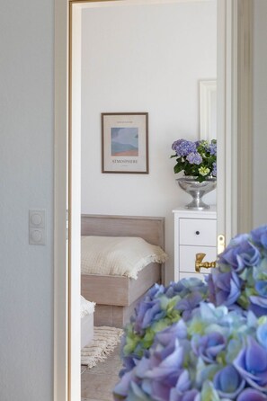 Ein Schlafzimmer mit einem Bett, einem Nachttisch mit Blumenvase, einem gerahmten Bild an der Wand und einer Tür zu einem anderen Raum.