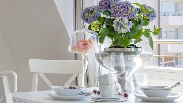 Uma mesa de jantar branca com uma cúpula de vidro que contém um doce, um vaso com flores e louça branca.