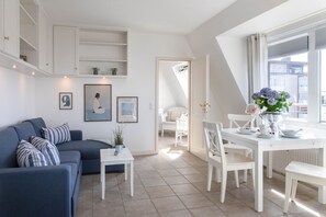 Ein modernes Wohnzimmer mit einem blauen Sofa, einem weißen Esstisch und Stühlen, einem kleinen Beistelltisch mit Pflanze und Blick durch ein Fenster.