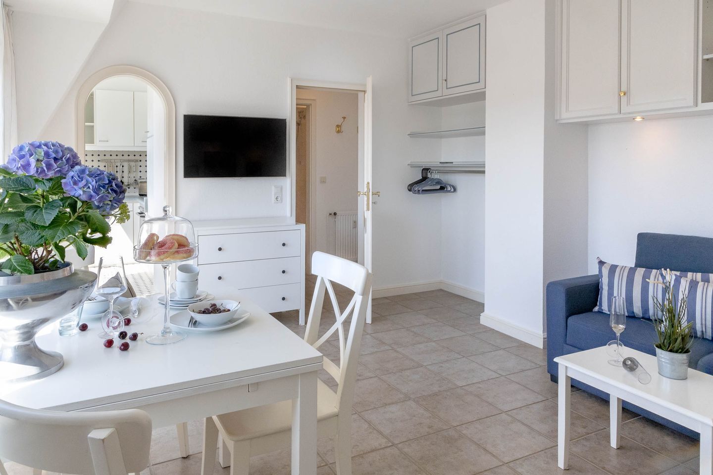 A modern living room with a dining area, white furniture, a flat-screen TV, and a blue sofa.