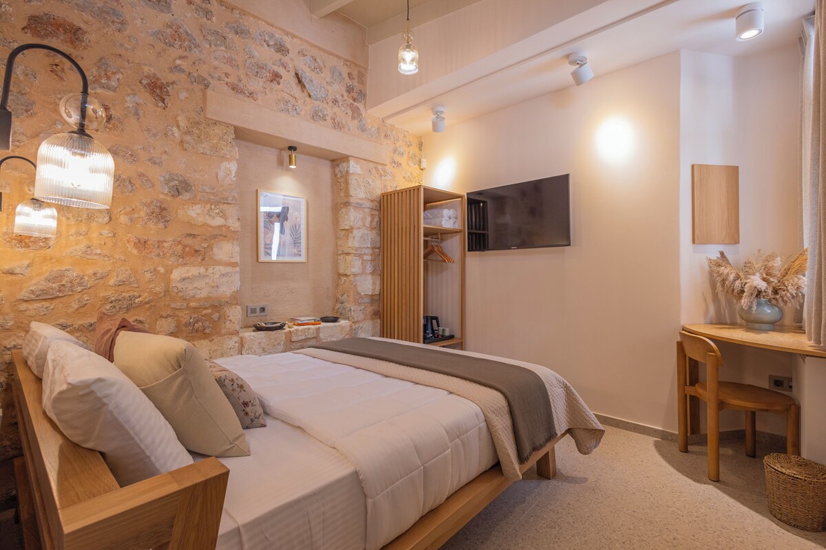 A bedroom with a stone wall, a bed with a wooden frame, a TV mounted on the wall, and a small table with a vase.