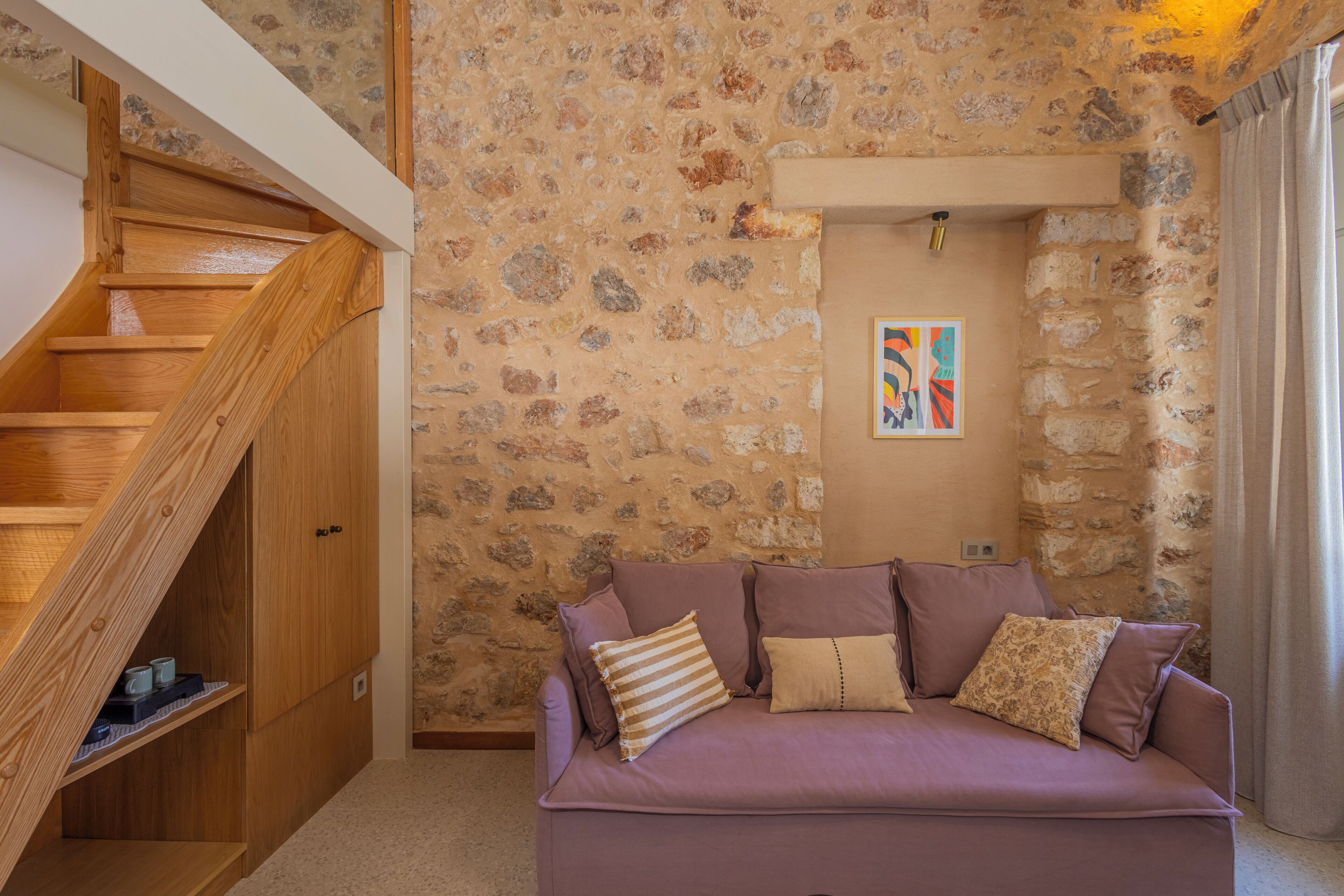 Une pièce avec un mur en pierre, un escalier en bois, un canapé violet et une petite table avec des tasses.