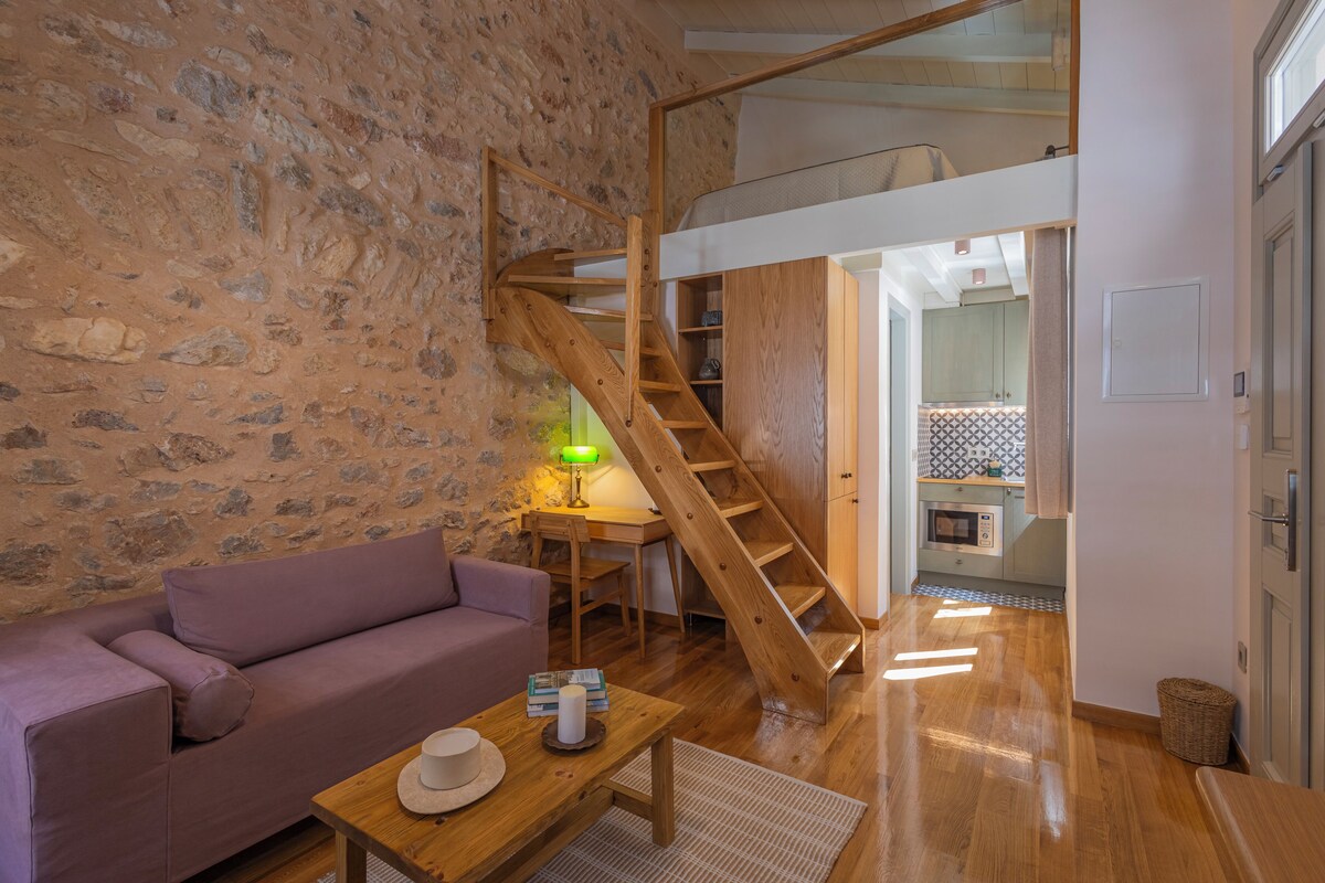 A cozy room with a stone wall, wooden staircase, a purple sofa, a wooden table with a hat, and a kitchen in the background.