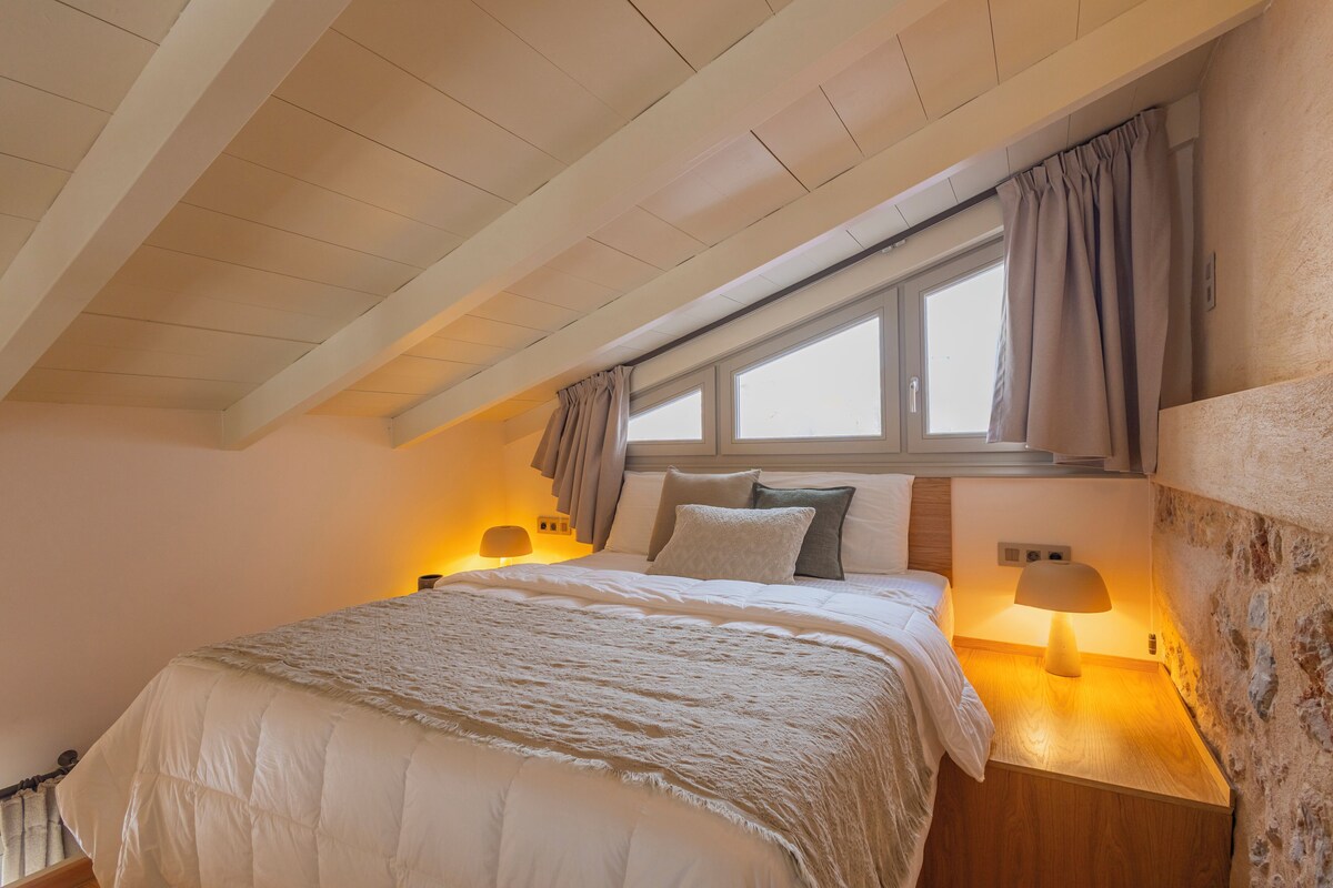 A bedroom with a sloped ceiling, a large bed with a white quilt, two bedside lamps, and a window with curtains.