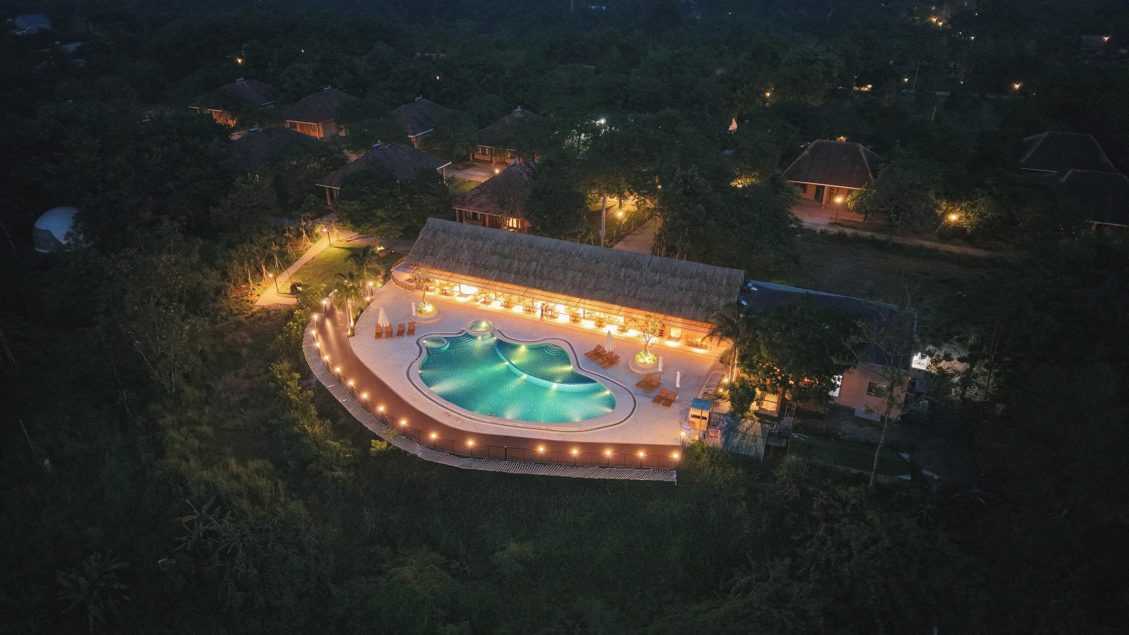 Vue aérienne d’un complexe hôtelier doté d’une piscine entourée de chaises longues et d’un espace couvert illuminé.