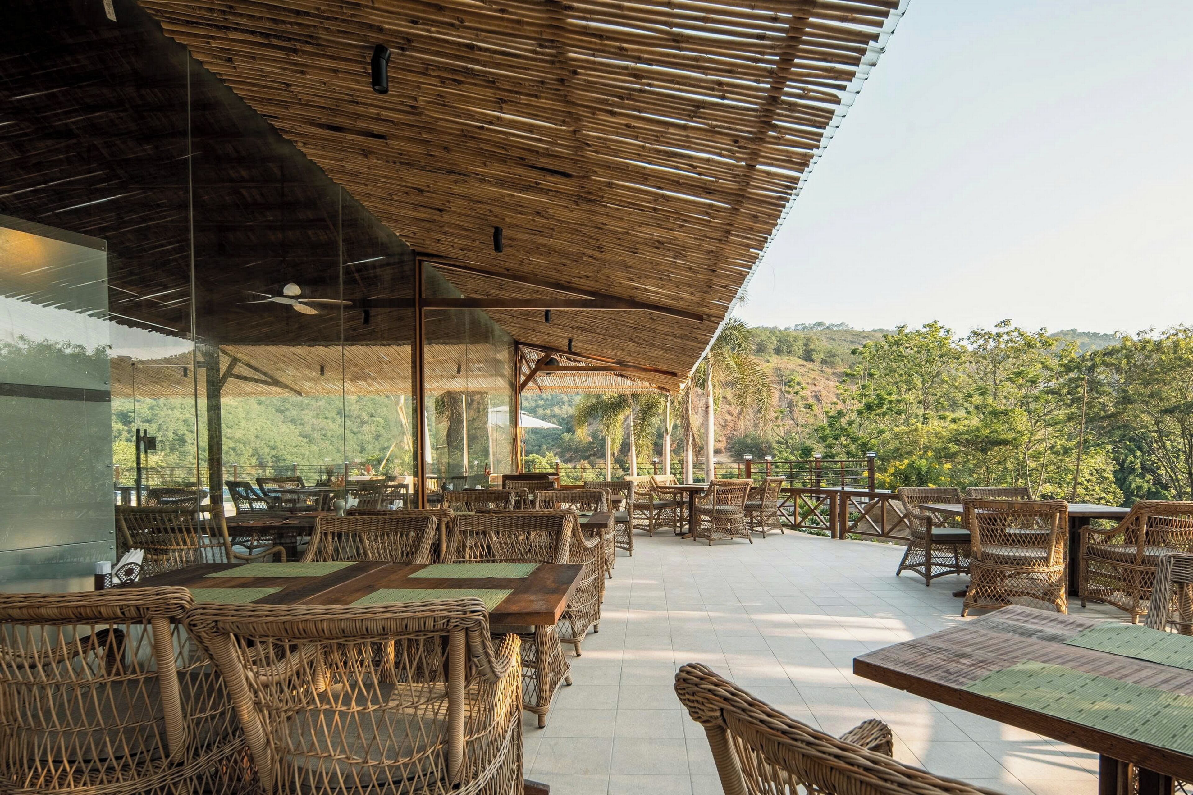 An outdoor restaurant with wicker furniture and a bamboo roof, overlooking a hilly landscape.